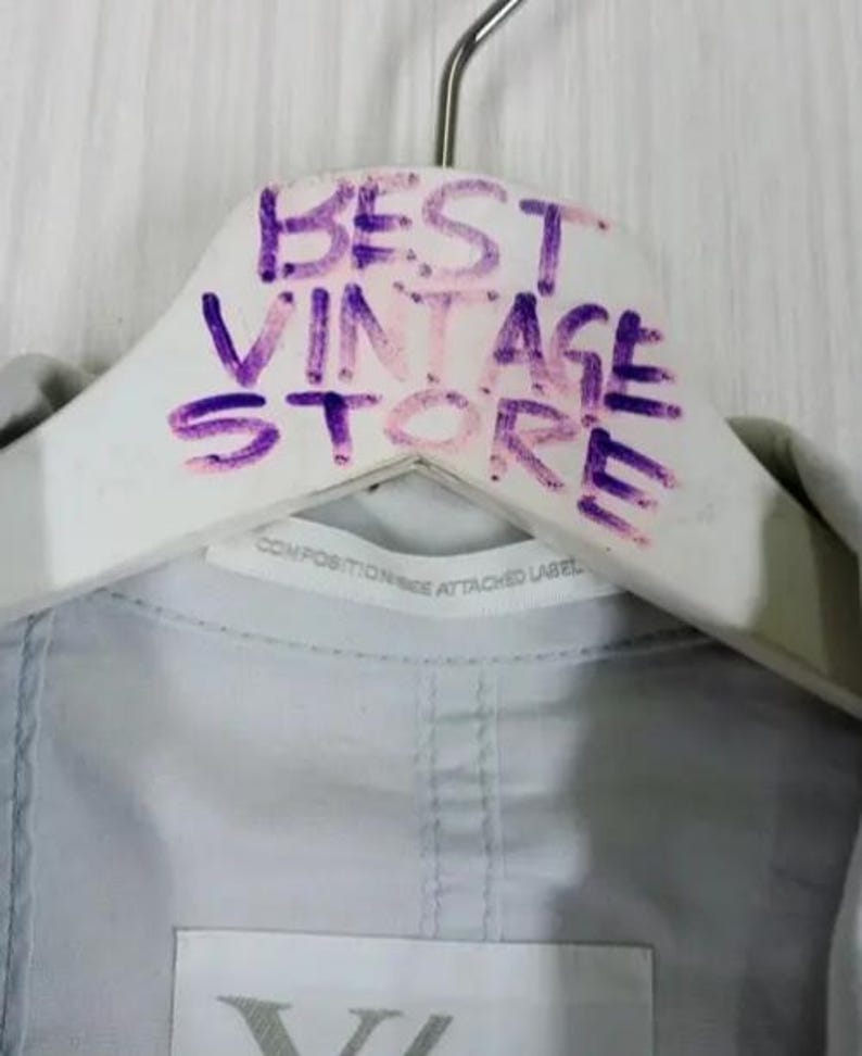 May include: A white wooden hanger with the words "BEST VINTAGE STORE" written in purple marker. The hanger is holding a light gray garment. A label with text is visible.