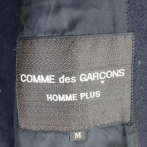 Pu&ograve; includere: Primo piano di un'etichetta nera con la scritta "COMME des GAR&Ccedil;ONS HOMME PLUS" in bianco. L'etichetta &egrave; cucita su un tessuto blu navy scuro. La taglia "M" &egrave; stampata su una piccola etichetta sottostante.