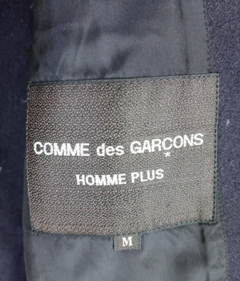 Pu&ograve; includere: Primo piano di un'etichetta nera con la scritta "COMME des GAR&Ccedil;ONS HOMME PLUS" in bianco. L'etichetta &egrave; cucita su un tessuto blu navy scuro. La taglia "M" &egrave; stampata su una piccola etichetta sottostante.