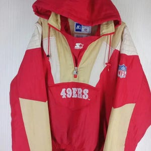 May include: Red, tan, and white hooded anorak jacket with the text "49ERS" across the front. The jacket has a half-zip closure, a drawstring hood, and the NFL logo on the right sleeve. The Starter brand logo is visible.