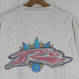 May include: A light gray sweatshirt with a graphic design on the back. The design features the letters "ZO" in red with a gray outline, surrounded by blue shapes and yellow flames. The sweatshirt is hanging on a wooden hanger.