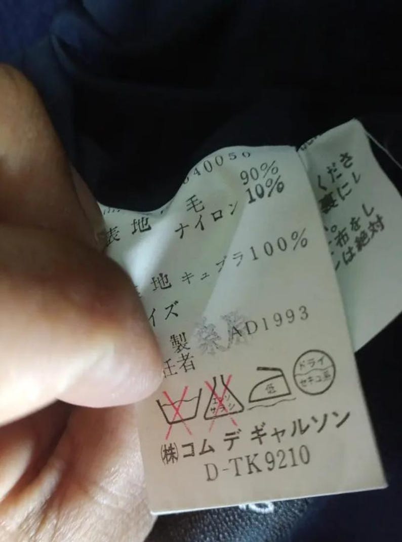 Pu&ograve; includere: Primo piano di un'etichetta di abbigliamento con testo giapponese, che include le percentuali di composizione del tessuto e le istruzioni per la cura. L'etichetta mostra anche il nome del marchio e il codice del prodotto.