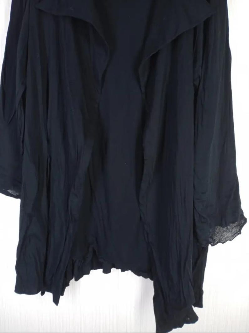 Op de afbeelding: Een lange, zwarte jas met een gedrapeerde kraag en lange mouwen. De stof lijkt licht van gewicht en heeft een licht gekreukte textuur. De zoom heeft een subtiele, gerimpelde rand, wat een vleugje detail aan het kledingstuk toevoegt.