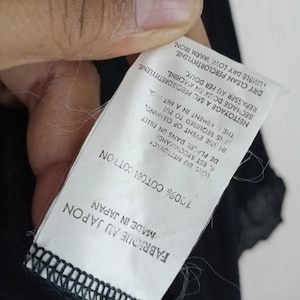 Op de afbeelding: Een wit label met zwarte tekst, vastgehouden door een hand, luidt "FABRIQUE AU JAPON MADE IN JAPAN 100% COTON COTTON". Extra tekst geeft reinigingsinstructies. Het label is bevestigd aan een zwart kledingstuk.