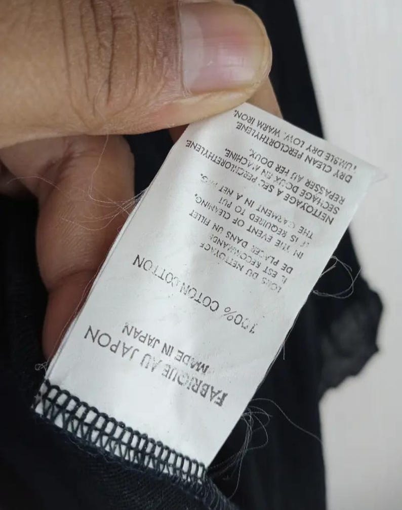 Op de afbeelding: Een wit label met zwarte tekst, vastgehouden door een hand, luidt "FABRIQUE AU JAPON MADE IN JAPAN 100% COTON COTTON". Extra tekst geeft reinigingsinstructies. Het label is bevestigd aan een zwart kledingstuk.