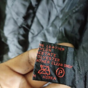 May include: Close-up of a black garment tag with red text. The tag reads "GENUINE LEATHER, NYLON, ACETATE, POLYESTER, LEATHER CLEAN ONLY" and includes a care symbol and the word "KOREA". The background is a quilted black fabric.