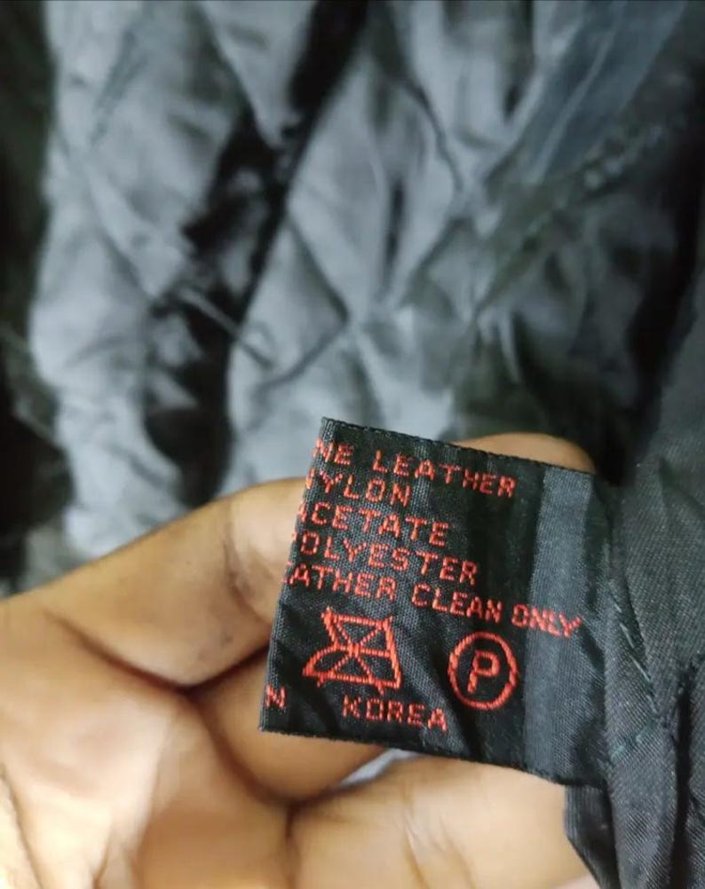 May include: Close-up of a black garment tag with red text. The tag reads "GENUINE LEATHER, NYLON, ACETATE, POLYESTER, LEATHER CLEAN ONLY" and includes a care symbol and the word "KOREA". The background is a quilted black fabric.