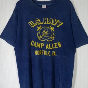 May include: Navy blue t-shirt with yellow text and graphics. The shirt features "U.S. NAVY" above an anchor emblem and "CAMP ALLEN NORFOLK, VA." below. The shirt is hanging on a wooden hanger.