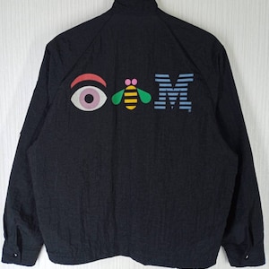 Könnte beinhalten: Schwarze Nylonjacke mit einem bunten Auge, einer Biene und einem "M"-Logo auf der Rückseite.
