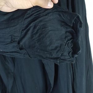 Op de afbeelding: Close-up van een zwart kledingstuk met lange mouwen. De mouw is bij de manchet gerimpeld, waardoor een getextureerd, gerimpeld effect ontstaat. De stof lijkt zacht en vloeiend, met zichtbare plooien en kreukels.