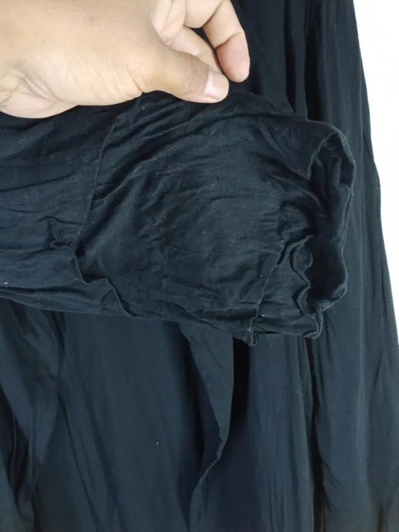 Op de afbeelding: Close-up van een zwart kledingstuk met lange mouwen. De mouw is bij de manchet gerimpeld, waardoor een getextureerd, gerimpeld effect ontstaat. De stof lijkt zacht en vloeiend, met zichtbare plooien en kreukels.