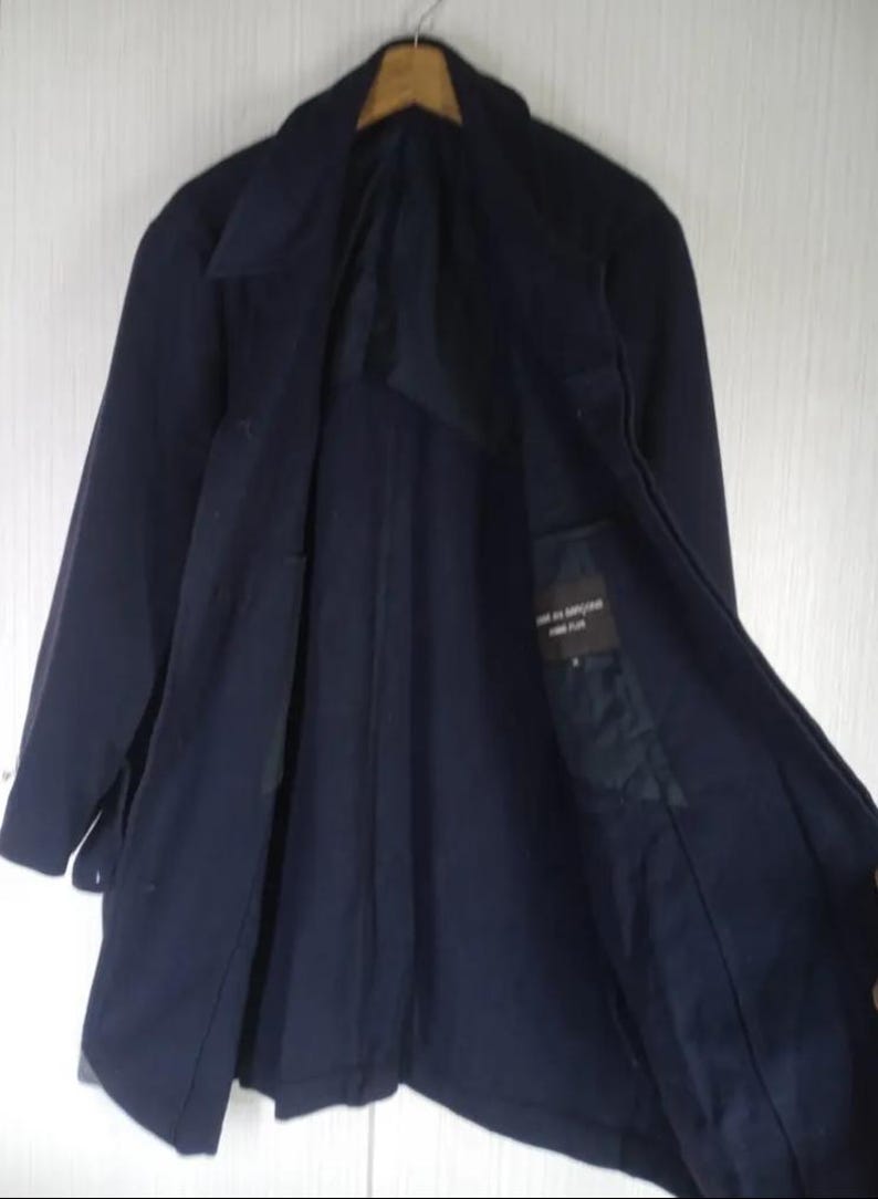 Pu&ograve; includere: Un cappotto blu navy dal design classico, appeso a una gruccia di legno. Il cappotto ha un colletto a revers, maniche lunghe e chiusura con bottoni. La fodera interna &egrave; visibile, con una piccola etichetta.