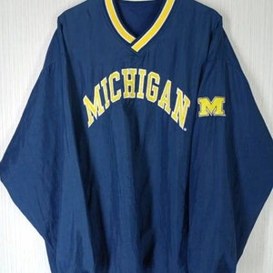 May include: A navy blue pullover jacket with a yellow V-neck collar and trim. The word "MICHIGAN" is arched across the chest in yellow lettering, with a yellow "M" logo on the right sleeve. The jacket is hanging on a wooden hanger.