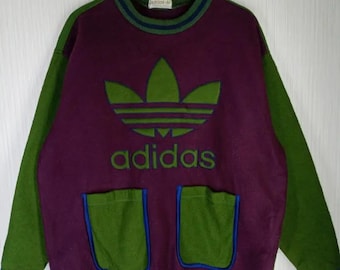 May include: A vintage Adidas sweatshirt in purple, green, and blue. The sweatshirt features the Adidas logo and text in green, with green sleeves and pockets. The collar and pockets are outlined in blue.