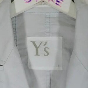 May include: A light gray jacket with a label that reads "Y's". The jacket is hanging on a white hanger with the text "VINTAGE STORE" written in purple marker. The jacket has a classic collar and a button-down front.