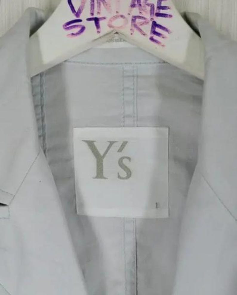 May include: A light gray jacket with a label that reads "Y's". The jacket is hanging on a white hanger with the text "VINTAGE STORE" written in purple marker. The jacket has a classic collar and a button-down front.