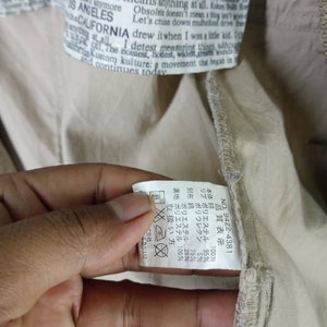 Peut inclure: Une &eacute;tiquette de v&ecirc;tement beige avec du texte noir et une &eacute;tiquette d'entretien blanche. L'&eacute;tiquette comprend le texte "NO. 9422-4381" et une liste de mat&eacute;riaux et de pourcentages. L'&eacute;tiquette d'entretien comprend des symboles pour le lavage, le repassage et le nettoyage &agrave; sec.