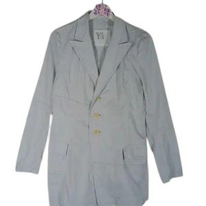 May include: A light gray blazer with a notched lapel, long sleeves, and three gold-colored buttons. The blazer has two front pockets and a split hem. The label reads "Y's". The blazer is hanging on a hanger.