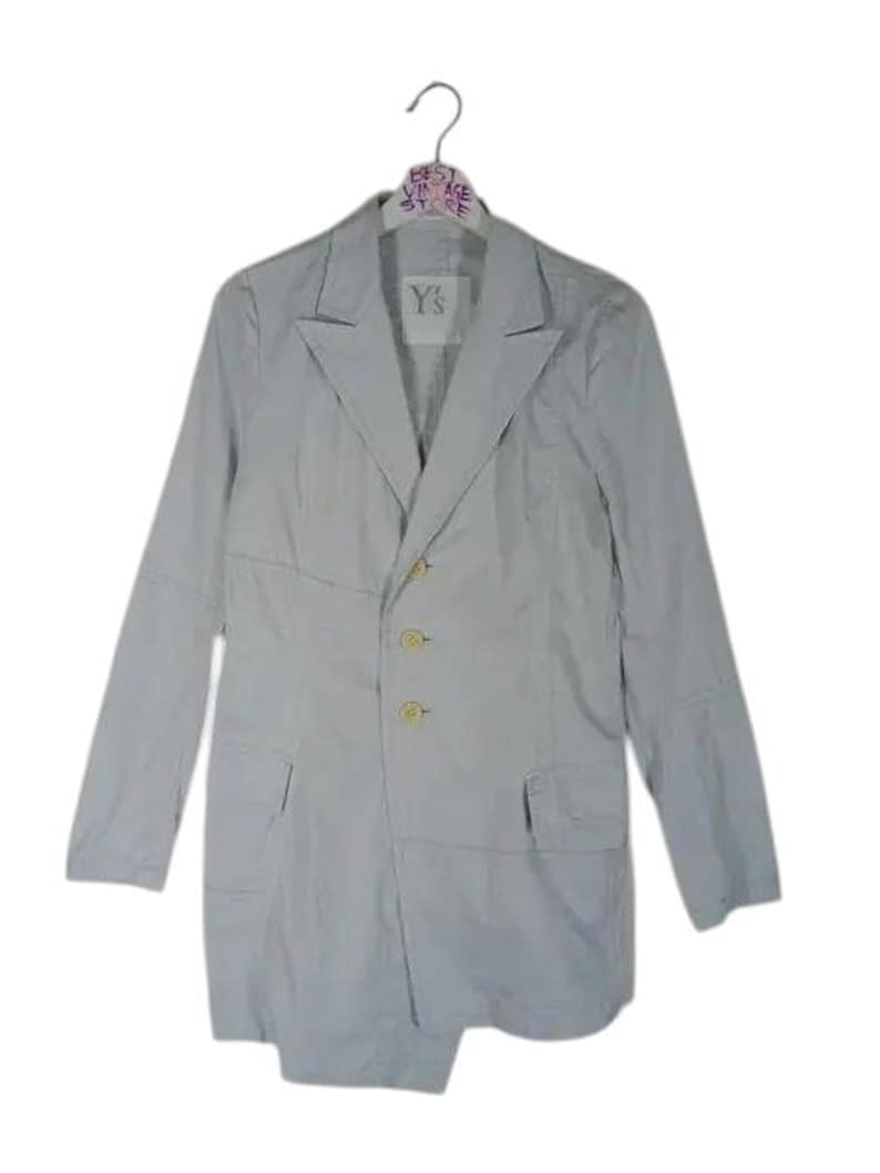 May include: A light gray blazer with a notched lapel, long sleeves, and three gold-colored buttons. The blazer has two front pockets and a split hem. The label reads "Y's". The blazer is hanging on a hanger.