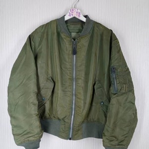 Puede incluir: Chaqueta bomber verde oliva con cremallera plateada de largo completo, dos bolsillos delanteros y cuello, puños y cintura de canalé. La chaqueta está colgada de una percha blanca. El texto en la percha dice "BEST VINTAGE STORE".