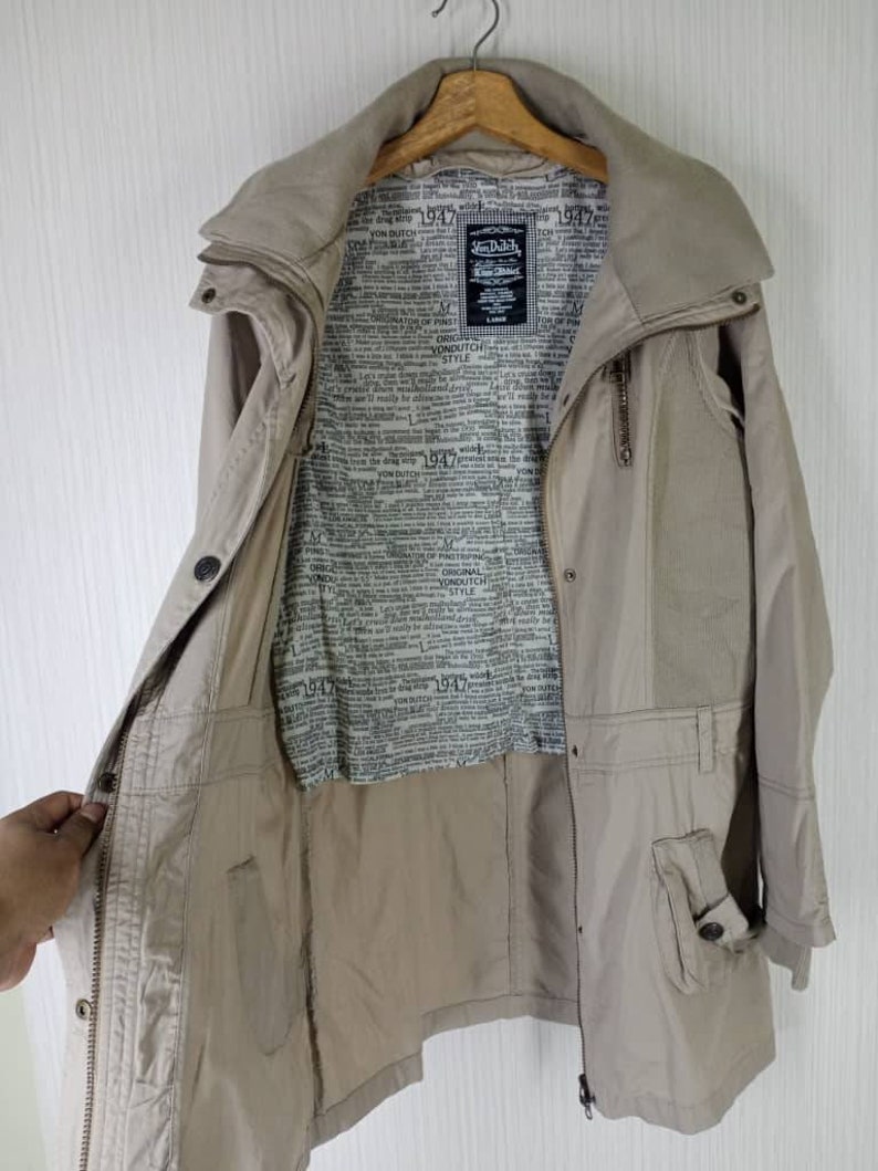 Peut inclure: Une veste beige &agrave; manches longues avec une fermeture &eacute;clair et une grande poche sur le c&ocirc;t&eacute; gauche. La veste est doubl&eacute;e d'un imprim&eacute; journal avec le texte "Von Dutch" et "Original Style" r&eacute;p&eacute;t&eacute; sur toute la surface.