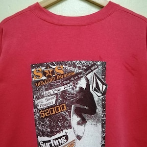 May include: Red crew neck sweatshirt featuring a graphic from the Volcom Airshow, with text including "July 29th, 1999" and "54th Street Newport". The graphic shows a surfer and the Vans logo.