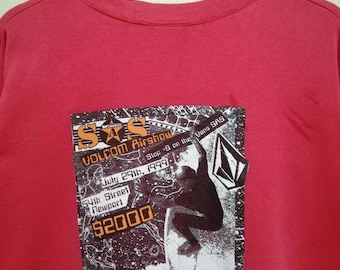 Può includere: Felpa rossa girocollo con grafica del Volcom Airshow, con testo come "July 29th, 1999" e "54th Street Newport". La grafica mostra un surfista e il logo Vans.
