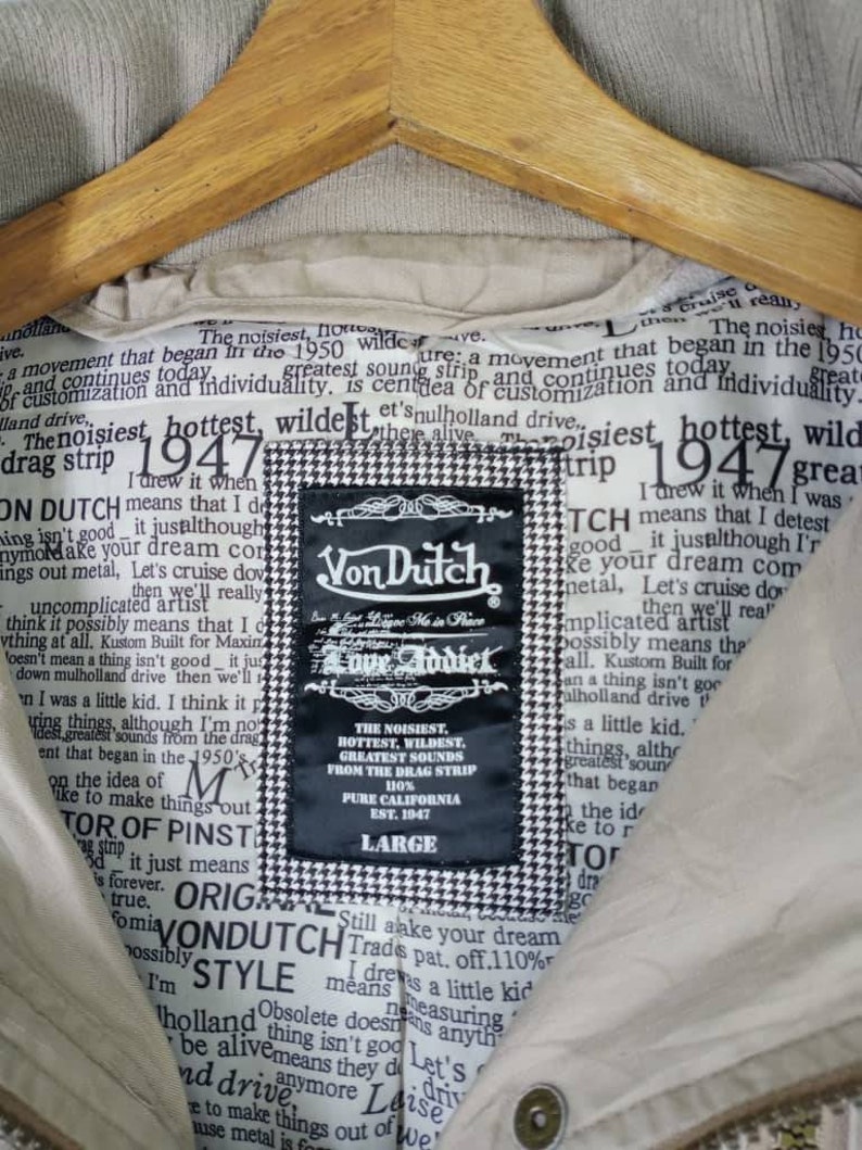 Peut inclure: Une veste beige avec un motif &agrave; carreaux noir et blanc. La veste porte une &eacute;tiquette qui dit "Von Dutch Love Addict" et comprend le texte "The noisiest, hottest, wildest, greatest sounds from the drag strip 110% Pure California Est. 1947".