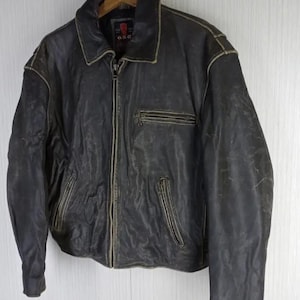 May include: A vintage, distressed black leather jacket with a zip-up front, collared neckline, and multiple pockets. The jacket shows signs of wear, with faded areas and a worn appearance. The jacket is hanging on a wooden hanger.