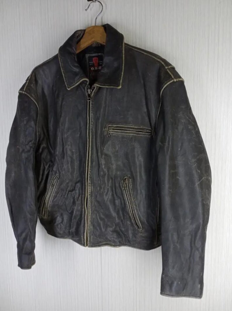 May include: A vintage, distressed black leather jacket with a zip-up front, collared neckline, and multiple pockets. The jacket shows signs of wear, with faded areas and a worn appearance. The jacket is hanging on a wooden hanger.