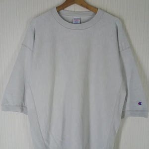 Peut inclure: Un sweat-shirt Champion gris clair à manches courtes, suspendu à un cintre en bois. Il présente un col rond, des manches courtes et le logo Champion sur la manche gauche. Le tissu semble doux et confortable.