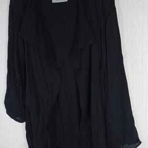 Op de afbeelding: Zwarte jas met lange mouwen en een gedrapeerde, open voorkant. De jas hangt aan een houten hanger. Een wit label met zwarte tekst is zichtbaar bij de halslijn. De stof lijkt licht en vloeiend.