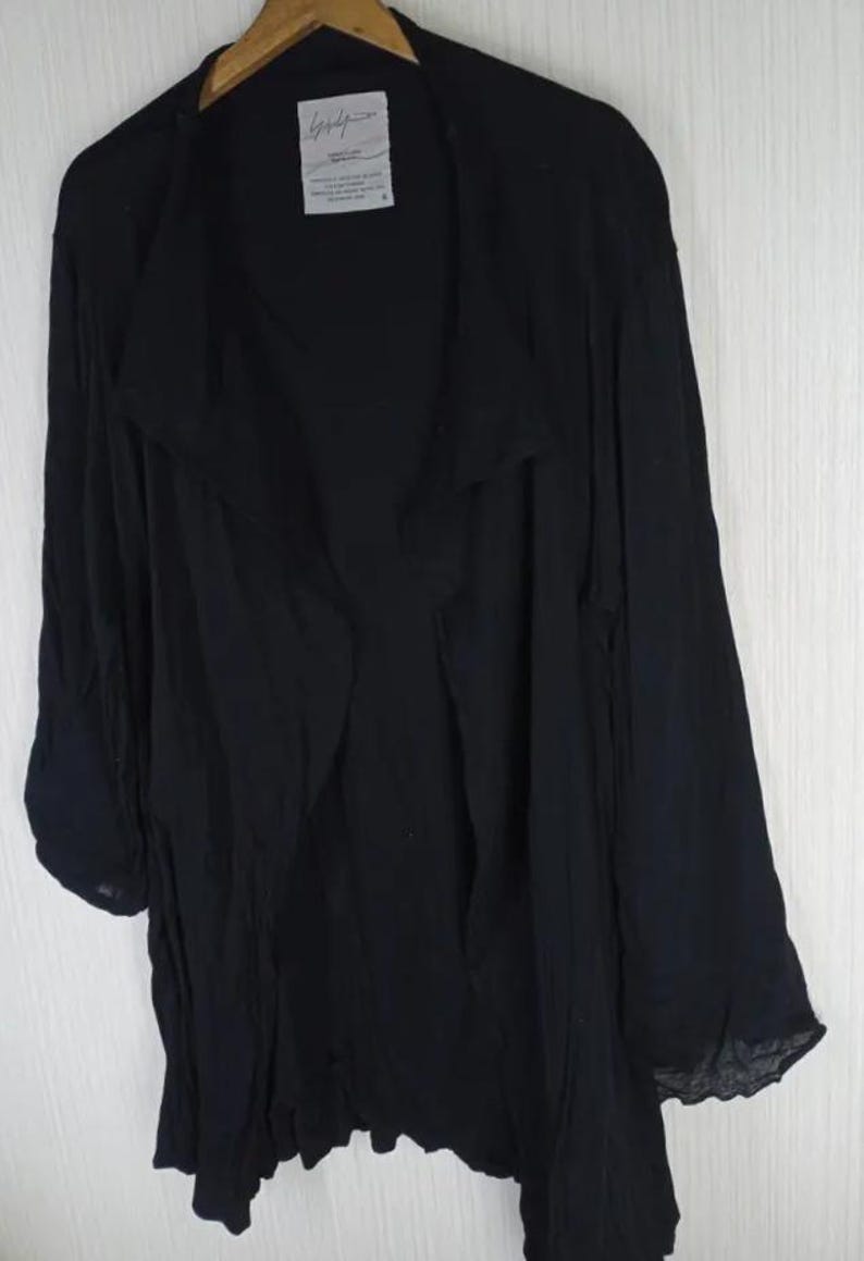 Op de afbeelding: Zwarte jas met lange mouwen en een gedrapeerde, open voorkant. De jas hangt aan een houten hanger. Een wit label met zwarte tekst is zichtbaar bij de halslijn. De stof lijkt licht en vloeiend.