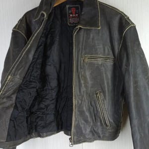 May include: A distressed brown leather jacket with a zip-up front, a collared neckline, and a black quilted lining. The jacket has two zippered pockets and a small label on the upper chest. The jacket is hanging on a wooden hanger.