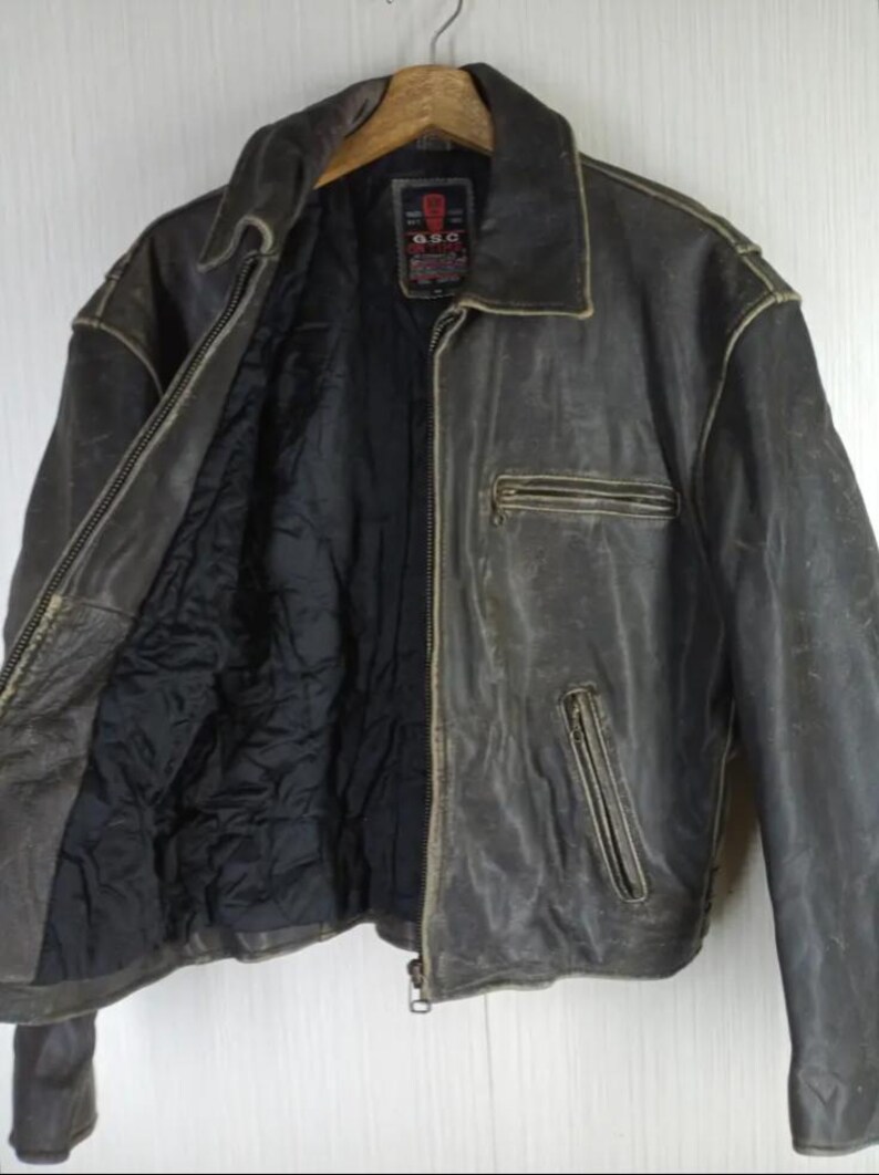 May include: A distressed brown leather jacket with a zip-up front, a collared neckline, and a black quilted lining. The jacket has two zippered pockets and a small label on the upper chest. The jacket is hanging on a wooden hanger.
