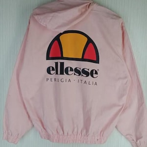 May include: A light pink jacket with a hood. The back of the jacket features the brand name "ellesse" in black, with a logo of a half-circle in yellow, red, and black. The text "PERUGIA - ITALIA" is below the brand name.