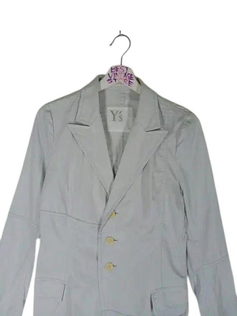 May include: A light gray blazer with three buttons and two pockets. The blazer is hanging on a hanger with the text "BEST VINTAGE STORE". The label on the blazer reads "Y's".