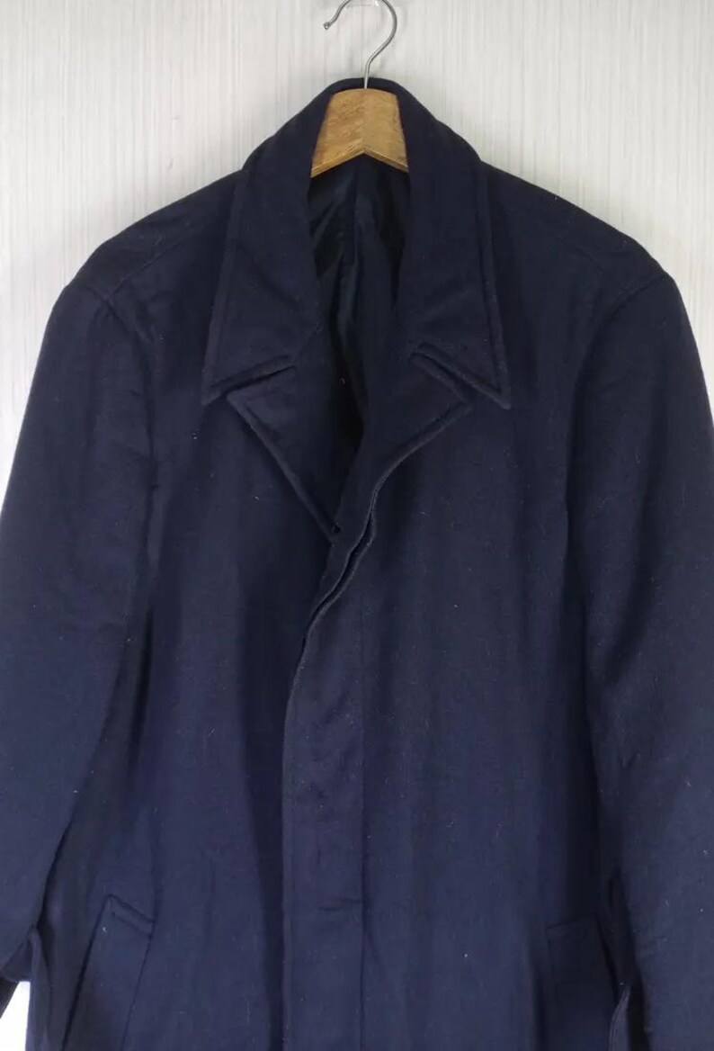 Pu&ograve; includere: Un cappotto blu navy in lana dal design classico. Il cappotto presenta un risvolto a lancia, una chiusura con bottoni e una lunghezza lunga. Il cappotto &egrave; appeso a un'appendiabiti di legno.