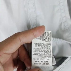 May include: Close-up of a white garment tag with text in English and French. The tag reads "MADE IN JAPAN" and "100% COTTON COTTON". Instructions include "HAND WASH" and "DO NOT BLEACH".