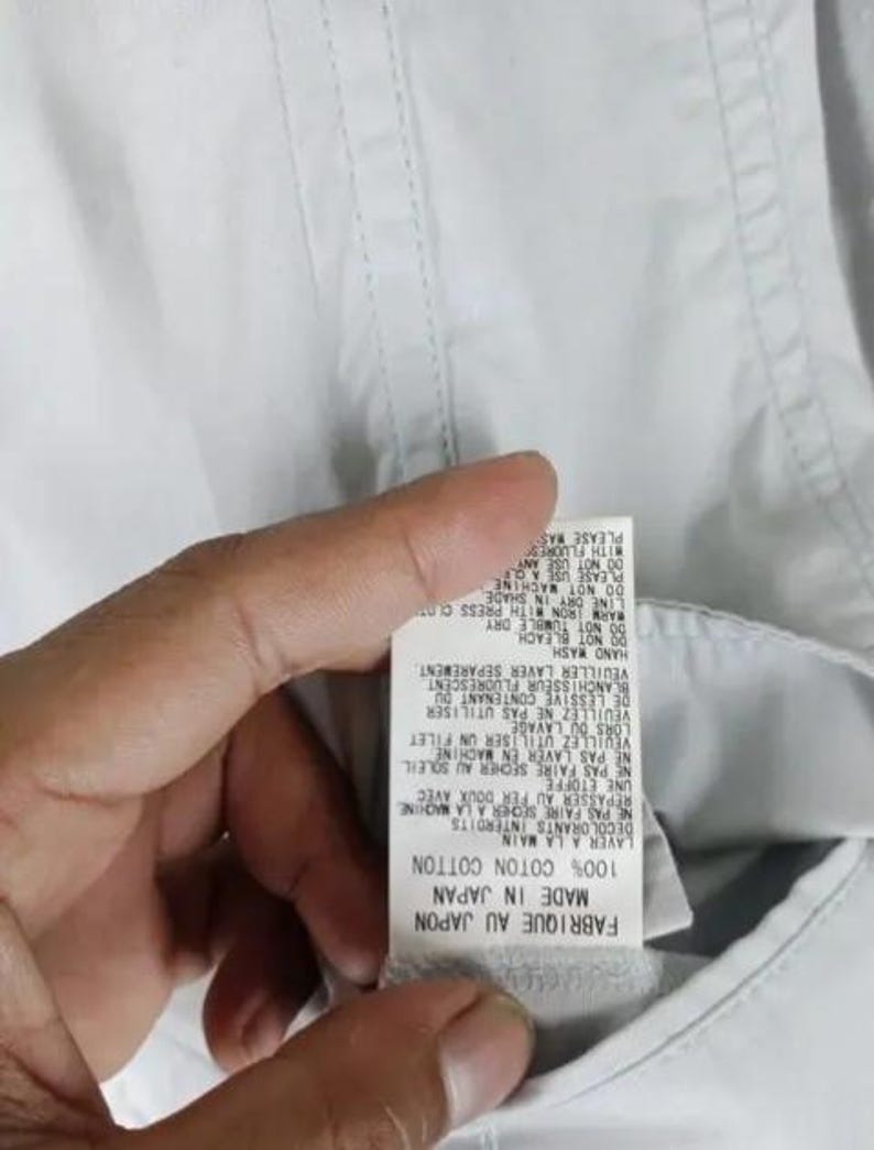 May include: Close-up of a white garment tag with text in English and French. The tag reads "MADE IN JAPAN" and "100% COTTON COTTON". Instructions include "HAND WASH" and "DO NOT BLEACH".