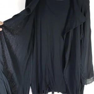 Op de afbeelding: Zwarte open vest met lange mouwen. Het kledingstuk heeft een gedrapeerd ontwerp en een label met Japanse tekst. De mouwen hebben een kanten rand aan de manchetten. De stof lijkt licht en vloeiend.