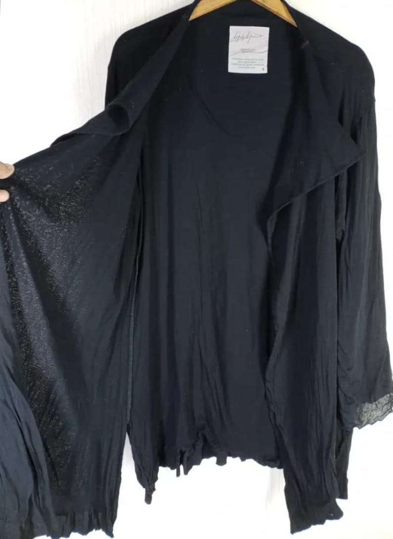 Op de afbeelding: Zwarte open vest met lange mouwen. Het kledingstuk heeft een gedrapeerd ontwerp en een label met Japanse tekst. De mouwen hebben een kanten rand aan de manchetten. De stof lijkt licht en vloeiend.