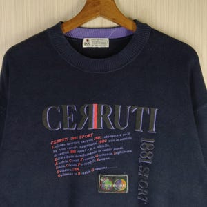 Könnte beinhalten: Marineblaues Cerruti 1881 Sport Sweatshirt. Das Sweatshirt zeigt den Markennamen in großen, mehrfarbigen Buchstaben auf der Brust, mit zusätzlichem Text und einem Logo darunter. Der Kragen ist lila und schwarz.
