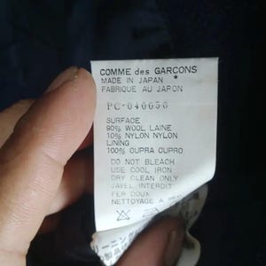 Pu&ograve; includere: Primo piano di un'etichetta di un capo Comme des Garcons. L'etichetta &egrave; bianca con testo nero, tra cui "MADE IN JAPAN" e dettagli sulla composizione del tessuto: 90% lana, 10% nylon e 100% cupro. Sono stampate anche le istruzioni per la cura.
