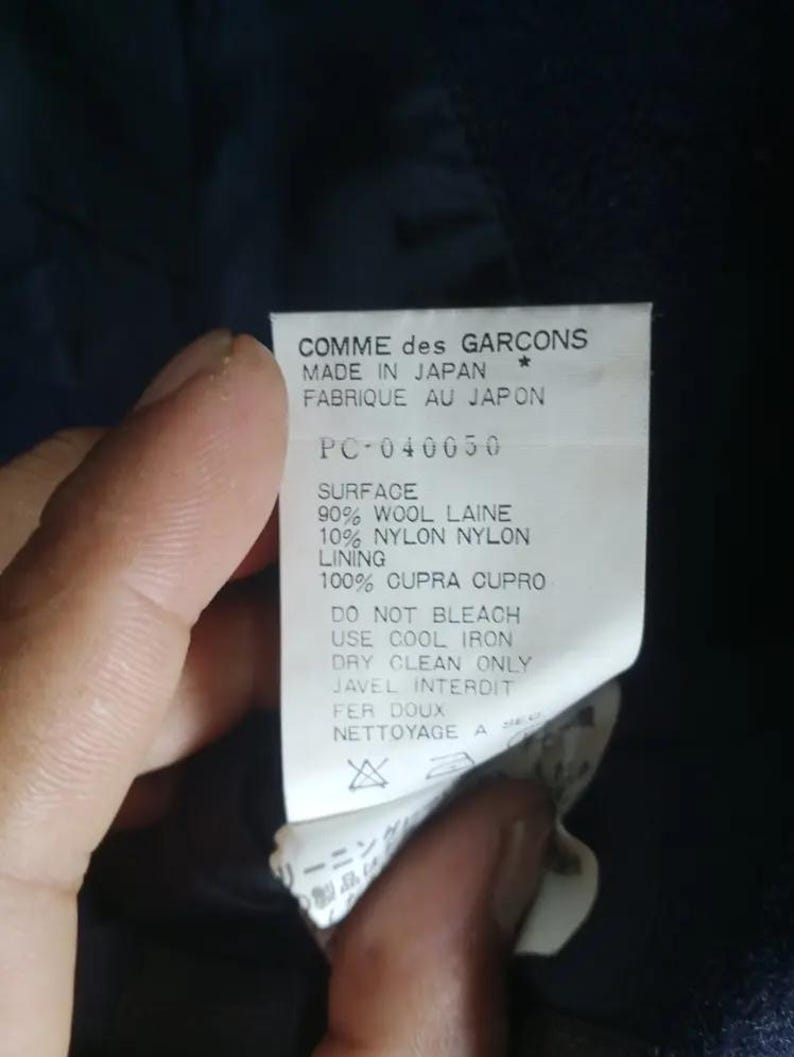 Pu&ograve; includere: Primo piano di un'etichetta di un capo Comme des Garcons. L'etichetta &egrave; bianca con testo nero, tra cui "MADE IN JAPAN" e dettagli sulla composizione del tessuto: 90% lana, 10% nylon e 100% cupro. Sono stampate anche le istruzioni per la cura.