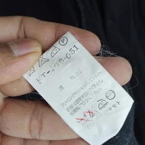 Op de afbeelding: Een wit kledinglabel met zwarte tekst en verzorgingssymbolen. Het label luidt "FT-106-051" en bevat wasvoorschriften. Het label wordt tegen een donkere stoffen achtergrond gehouden.