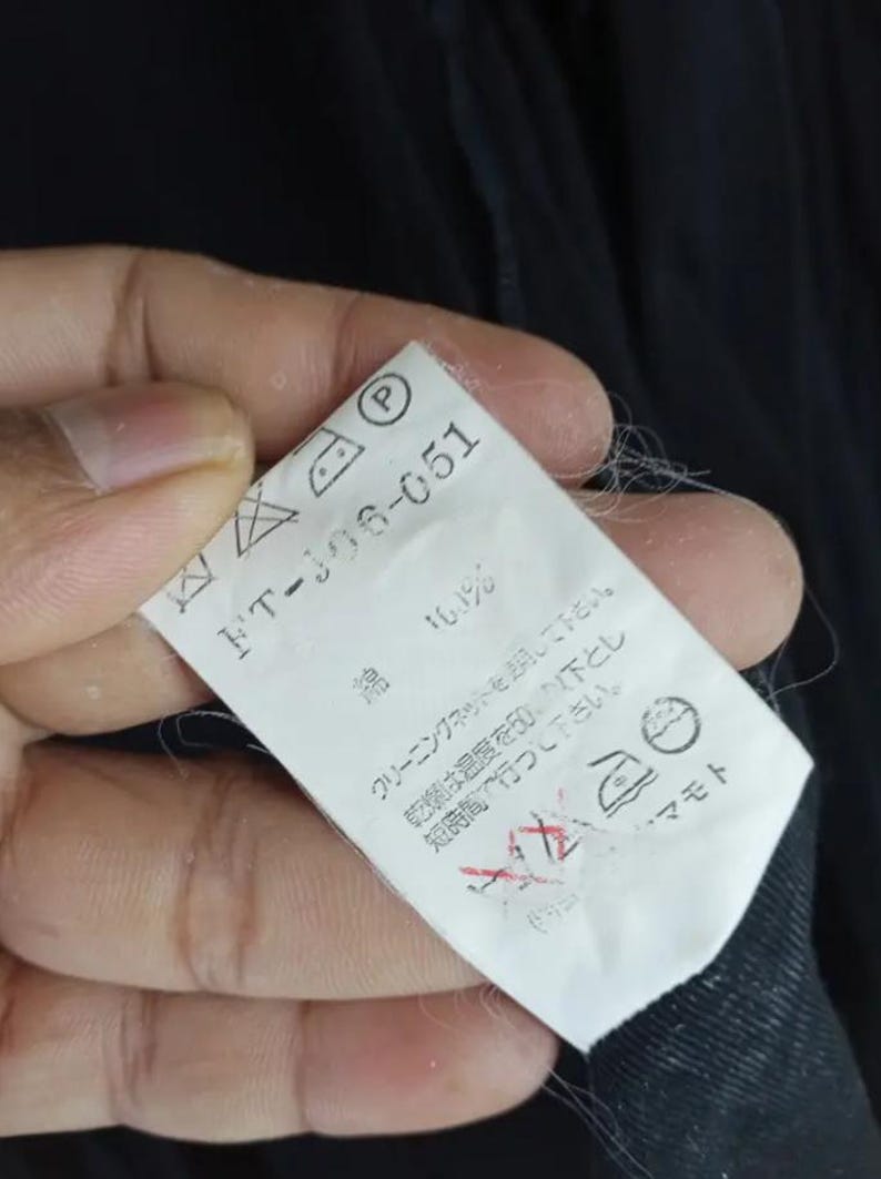 Op de afbeelding: Een wit kledinglabel met zwarte tekst en verzorgingssymbolen. Het label luidt "FT-106-051" en bevat wasvoorschriften. Het label wordt tegen een donkere stoffen achtergrond gehouden.