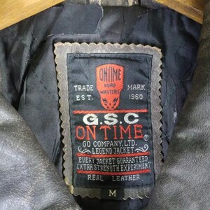 May include: Close-up of a vintage leather jacket's interior, showcasing a black label with red and white text. The label reads "ON TIME" and "REAL LEATHER." The jacket is a weathered gray color, with a size "M" indicated on the label.