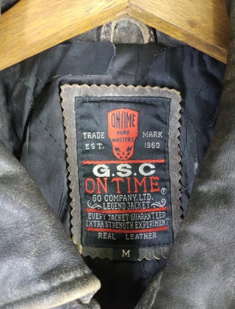 May include: Close-up of a vintage leather jacket's interior, showcasing a black label with red and white text. The label reads "ON TIME" and "REAL LEATHER." The jacket is a weathered gray color, with a size "M" indicated on the label.