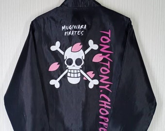 以下が含まれることがあります： 黒色のジャケット。スカルとクロスボーンのグラフィック、ピンクのアクセント、そして「Mugiwara Pirates」と「Tony Tony. Chopper」の文字が特徴です。 ジャケットはハンガーに掛けられています。